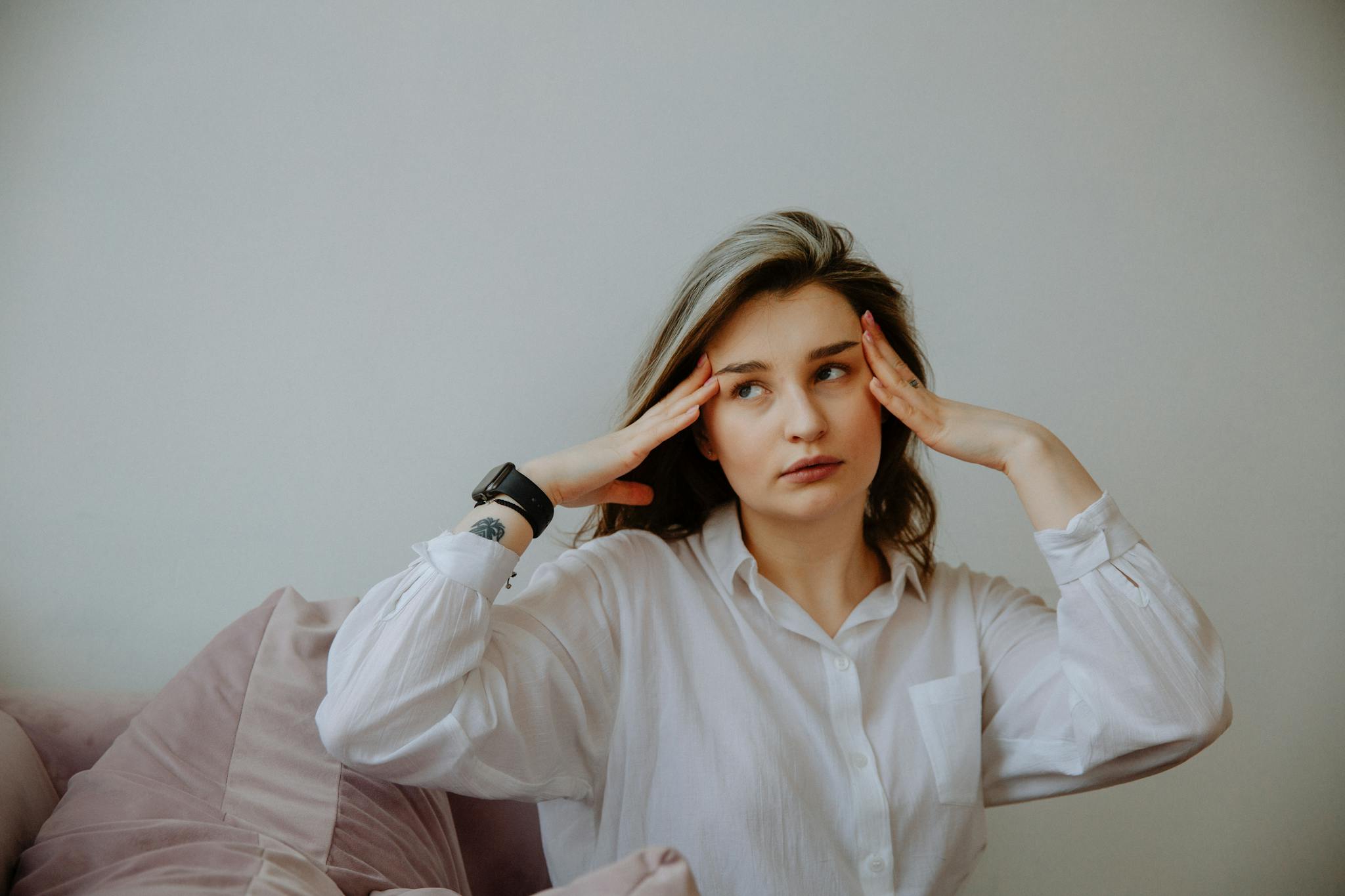 Woman in white shirt looking stressed, touching her temples indoors.